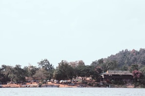 A scenic lakeside view featuring a small house or cabin nestled among lush trees and foliage. The area is surrounded by a dense forest, and a few parked cars are visible near the shore. The calm lake occupies the foreground, with its clear, serene water. The sky is light, adding to the tranquil atmosphere.