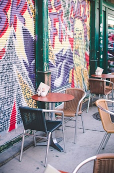 Street art featuring bold graffiti covers the exterior wall beside an outdoor café setting. Several tables and chairs, crafted from metal and wicker, are placed along the sidewalk. A menu and a small holder sit on one of the tables, indicating a ready setup for visitors.