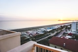 A coastal landscape at sunset with the sun setting on the horizon over the sea. The foreground features buildings with terracotta roofs and a balcony railing. Beyond, a beach with beach huts lined up can be seen, and a vast expanse of calm water stretches towards the horizon. A road runs parallel to the beach lined with palm trees.