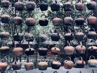 A collection of rustic, clay teapots hanging in rows against a background of lush greenery and traditional architecture. The teapots exhibit a variety of earthy tones, showing signs of age and use.