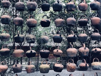 A collection of rustic, clay teapots hanging in rows against a background of lush greenery and traditional architecture. The teapots exhibit a variety of earthy tones, showing signs of age and use.