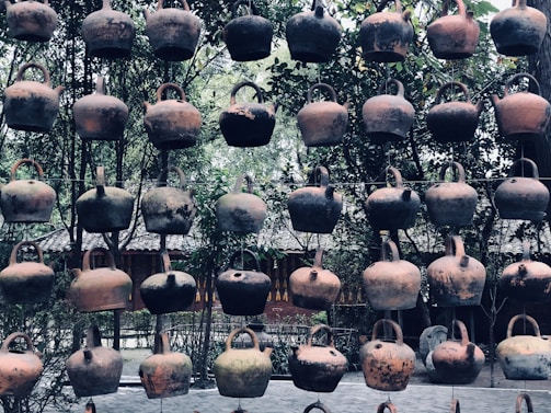 A collection of rustic, clay teapots hanging in rows against a background of lush greenery and traditional architecture. The teapots exhibit a variety of earthy tones, showing signs of age and use.