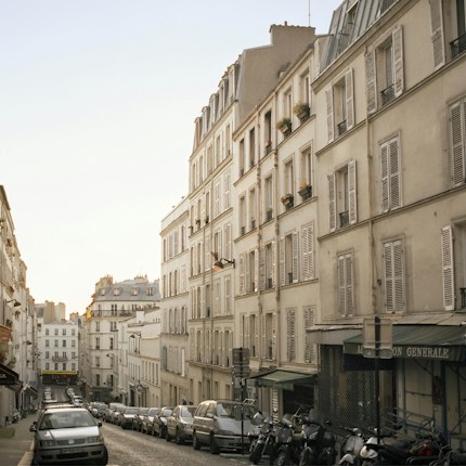 A quaint, narrow street lined with classic Parisian buildings featuring white facades and shuttered windows. The cobblestone road is lined with parked cars and motorcycles, and the glow of early evening light gives the street a warm, serene ambiance.