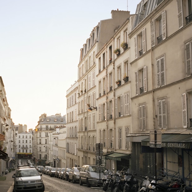 A quaint, narrow street lined with classic Parisian buildings featuring white facades and shuttered windows. The cobblestone road is lined with parked cars and motorcycles, and the glow of early evening light gives the street a warm, serene ambiance.