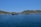 A serene coastal landscape featuring a sprawling fish farm on a calm blue sea. Hills with sparse vegetation rise in the background under a clear blue sky. The fish enclosures consist of various floating structures in the water.