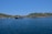 A serene coastal landscape featuring a sprawling fish farm on a calm blue sea. Hills with sparse vegetation rise in the background under a clear blue sky. The fish enclosures consist of various floating structures in the water.