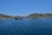 A serene coastal landscape featuring a sprawling fish farm on a calm blue sea. Hills with sparse vegetation rise in the background under a clear blue sky. The fish enclosures consist of various floating structures in the water.