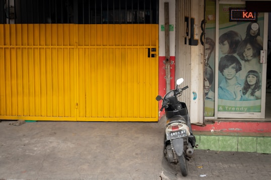 A yellow metal gate is positioned next to a small store entrance with a poster featuring various hairstyles. A black scooter with a visible license plate is parked near the entrance, resting on a concrete surface.