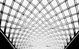 An intricate geometric pattern of intersecting metal and glass beams forms a large dome structure. The design is symmetrical and visually captivating, with the framework creating a lattice of diamonds and triangles.