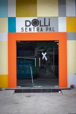 A modern storefront entrance with a colorful square-patterned facade. The entrance is framed by a bold orange border, and the doors are made of glass with silver handles. The interior is partially visible through the glass, including a blue object that appears to be discarded items. The word 'Dolli' is prominently displayed above the entrance in black, stylized text.