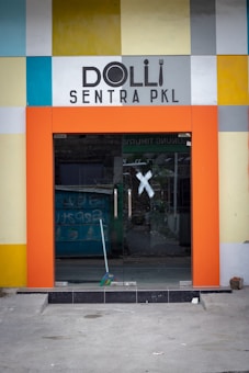 A modern storefront entrance with a colorful square-patterned facade. The entrance is framed by a bold orange border, and the doors are made of glass with silver handles. The interior is partially visible through the glass, including a blue object that appears to be discarded items. The word 'Dolli' is prominently displayed above the entrance in black, stylized text.
