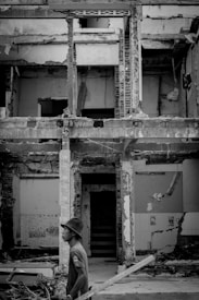 A man wearing a hat and holding a wooden plank stands in front of a partially demolished building. The structure shows exposed bricks and beams, with remnants of paint and tiles. Debris and construction materials are scattered around the area.
