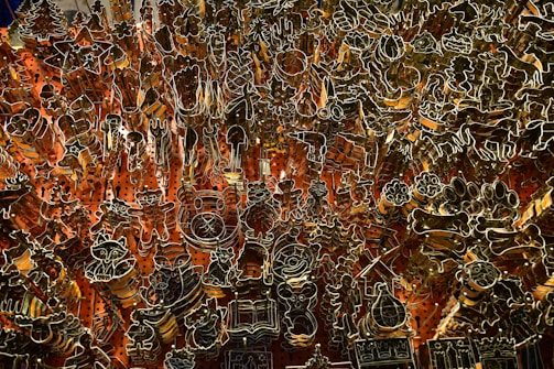 An intricate array of metal cookie cutters is organized against a pegboard with red-orange backlighting. The cutters are various shapes including animals, stars, trees, and other festive designs.