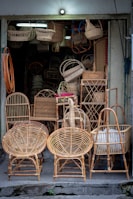 A small shop displays a variety of wicker furniture and items. Two large wicker chairs are prominently placed in the foreground, showcasing their intricate woven patterns. Behind them are baskets, stools, and other woven goods, filling the entrance area. The setting is dimly lit with natural colors highlighting the craftsmanship.