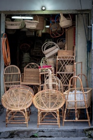 A small shop displays a variety of wicker furniture and items. Two large wicker chairs are prominently placed in the foreground, showcasing their intricate woven patterns. Behind them are baskets, stools, and other woven goods, filling the entrance area. The setting is dimly lit with natural colors highlighting the craftsmanship.