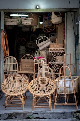 A small shop displays a variety of wicker furniture and items. Two large wicker chairs are prominently placed in the foreground, showcasing their intricate woven patterns. Behind them are baskets, stools, and other woven goods, filling the entrance area. The setting is dimly lit with natural colors highlighting the craftsmanship.