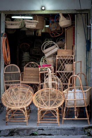 A small shop displays a variety of wicker furniture and items. Two large wicker chairs are prominently placed in the foreground, showcasing their intricate woven patterns. Behind them are baskets, stools, and other woven goods, filling the entrance area. The setting is dimly lit with natural colors highlighting the craftsmanship.
