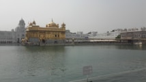 A golden temple sits majestically on the water, surrounded by serene reflections and a backdrop of white and gray structures. The temple's intricate architecture is highlighted by its gilded surfaces, domes, and ornate details. The surrounding water provides a tranquil atmosphere, enhancing the view of the temple.