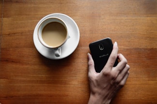 A close-up of hands holding a smartphone and a coffee cup on a wooden table.