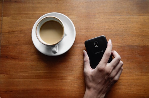 A close-up of hands holding a smartphone and a coffee cup on a wooden table.