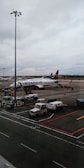 An airport scene featuring a Ryanair airplane parked on the tarmac with boarding stairs and several service vehicles nearby. The sky is overcast, and there is a large light pole in the foreground.