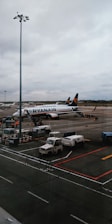 An airport scene featuring a Ryanair airplane parked on the tarmac with boarding stairs and several service vehicles nearby. The sky is overcast, and there is a large light pole in the foreground.