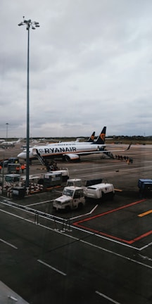 An airport scene featuring a Ryanair airplane parked on the tarmac with boarding stairs and several service vehicles nearby. The sky is overcast, and there is a large light pole in the foreground.