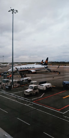 An airport scene featuring a Ryanair airplane parked on the tarmac with boarding stairs and several service vehicles nearby. The sky is overcast, and there is a large light pole in the foreground.