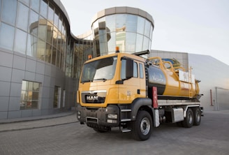 A large orange industrial truck with a tanker attached is parked in front of a modern glass building with reflective, curved windows. The building features a futuristic design with a sleek facade.