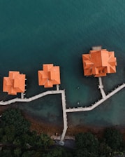 Aerial view showing a group of orange-roofed buildings on stilts connected by a series of pathways over an expanse of blue-green water. The pathways and buildings appear to be part of a coastal resort or settlement. Lush green vegetation lines the shore, contrasting with the clear water surrounding the structures.