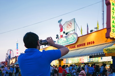 A person wearing a blue shirt photographs a crowded scene at a bustling outdoor event. Prominent signage for 'Nathan's' with a classic, retro design and an anniversary mention are visible, alongside yellow and red flags atop the building. A variety of people gather around, engaging with the festive atmosphere, while a distant amusement park ride adds to the scene's vibrancy.