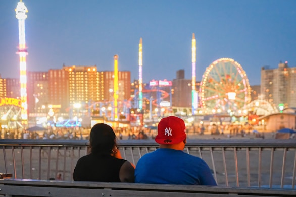 man and woman sitting on a waterfront bench looking at Coney Island at night
