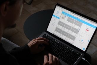 A person is using a laptop, typing with both hands on the keyboard. The screen displays a website layout with a grid of images and text panels.