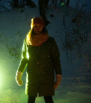 A person stands outdoors in a snowy environment, illuminated by a greenish light. The individual is wearing a warm coat, scarf, hat, and mittens, suggesting cold weather. Snowflakes or particles are visible in the air, enhancing the wintry atmosphere.