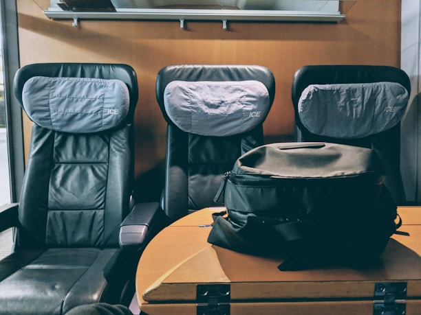 Three black leather passenger seats are arranged side by side with gray headrest covers labeled 'ICE'. In front of the seats, there is a wooden table with a black backpack resting on top.