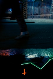 A blurred figure walks past a wet pavement reflecting neon lights, with a brick wall and metal gate in the background. The reflection creates an abstract scene with vibrant colors including orange and green.