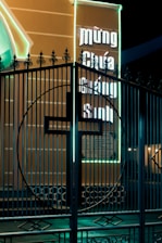 A decorative metal gate with an intricate design is shown in front of an illuminated billboard. The billboard has neon lights and contains text in a non-English script. The neon glows brightly against the dark night, casting a vivid contrast.