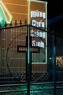 A decorative metal gate with an intricate design is shown in front of an illuminated billboard. The billboard has neon lights and contains text in a non-English script. The neon glows brightly against the dark night, casting a vivid contrast.