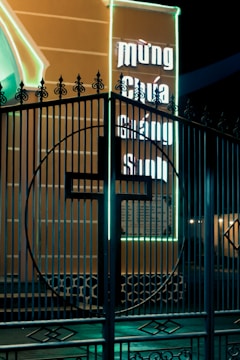 A decorative metal gate with an intricate design is shown in front of an illuminated billboard. The billboard has neon lights and contains text in a non-English script. The neon glows brightly against the dark night, casting a vivid contrast.