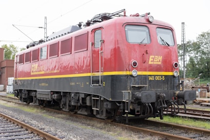 A red and black electric locomotive is stationed on railway tracks. It has a yellow stripe running along the side and the letters EBM are prominently displayed on the side and front. The locomotive is surrounded by railway infrastructure, including power lines and signal posts, and there are industrial buildings and trees in the background.
