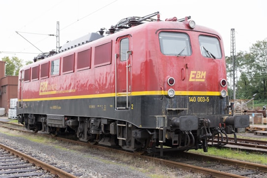 A red and black electric locomotive is stationed on railway tracks. It has a yellow stripe running along the side and the letters EBM are prominently displayed on the side and front. The locomotive is surrounded by railway infrastructure, including power lines and signal posts, and there are industrial buildings and trees in the background.