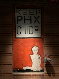A mural on a brick wall featuring a rectangular area with a primarily orange background. At the top, it has the text 'KEEP PHX CHIDO' in bold black letters, with a small shirt or smiley motif next to the 'O'. Below the text, there is an image of a stylized girl in white with a small smile, hair tied back, and what appears to be a wreath or headband. To the right of the mural, there is a metal AUTO SPKR fire department connection.