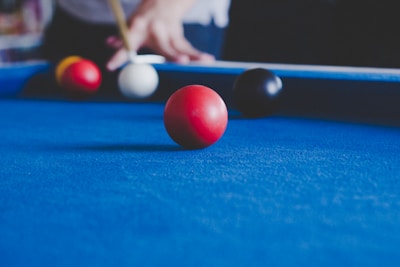 A close-up view of a pool table with vibrant blue felt. A red billiard ball is prominently in focus, while other balls, including yellow, white, and black, are positioned further back on the table. A person is seen in the background, holding a pool cue, poised for a shot.