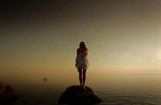 A person stands alone on a rock, gazing into a vast, calm body of water under a hazy sky. They are wearing a loose, long-sleeved garment with text on the back, and the scene suggests solitude and introspection.