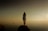 A person stands alone on a rock, gazing into a vast, calm body of water under a hazy sky. They are wearing a loose, long-sleeved garment with text on the back, and the scene suggests solitude and introspection.