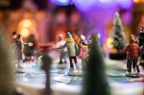 A miniature scene of people ice skating, featuring small figurines wearing winter clothing. Trees are visible around the skating rink, and the background is softly lit with warm, blurry lights, creating a cozy, festive atmosphere.