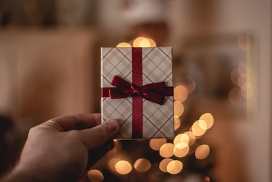 A hand is holding a small, rectangular gift box wrapped in plaid paper with a red ribbon tied in a bow. The background is blurred, featuring warm bokeh lights that suggest a festive or holiday setting.