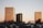 Three tall buildings with reflective glass facades are set against a clear sky during sunset. Each building has the name of a company on its top. There is a line of parked cars in the foreground, and a few trees around the parking lot area.