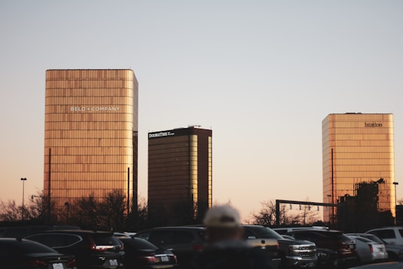 Three tall buildings with reflective glass facades are set against a clear sky during sunset. Each building has the name of a company on its top. There is a line of parked cars in the foreground, and a few trees around the parking lot area.