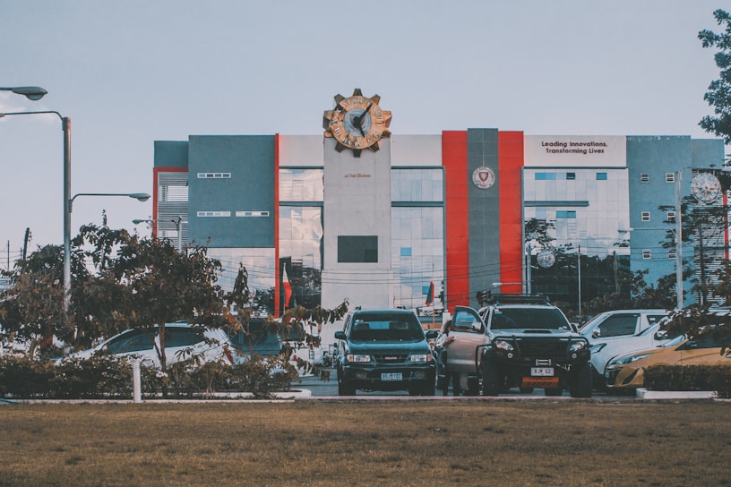 A modern building with a large emblem at the top is visible; it features gray, red, and blue tones. In front of the building, there is a row of parked cars and some greenery, with a lawn in the foreground. The building has a slogan about innovation visible on its facade.
